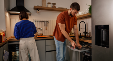 a woman and man cleaning the kitchen and he is loading a dishwasher